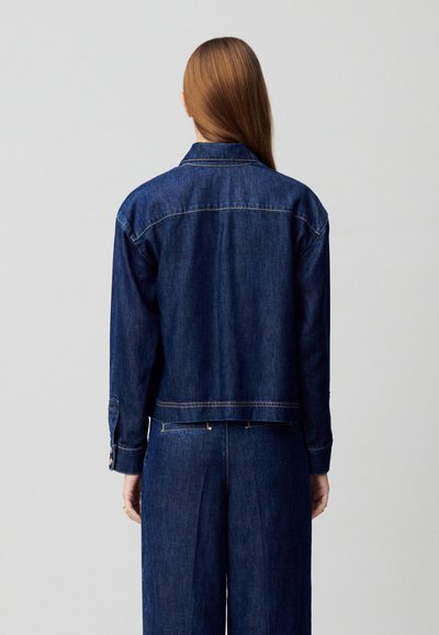 Chaqueta de denim de color azul oscuro con cuello, mangas largas y puños abotonados. La parte trasera tiene costuras mínimas y un diseño recortado.