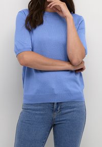 Donna con lunghi capelli scuri, che indossa un maglione blu a maniche corte e jeans blu, con un braccio incrociato e una mano vicino alla bocca.