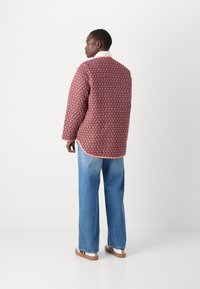 Röd mönstrad jacka med en ljusgrå krage, kombinerad med ljusblå vida jeans och vita skor med bruna sulor. Bakvy.