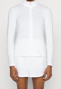 White long-sleeve zip-up top with high collar, made from smooth, lightweight fabric. Features subtle logo and contrasting zipper detail.