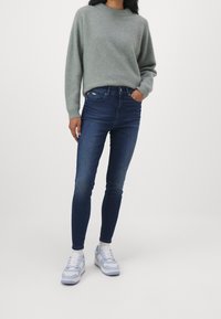Suéter de punto gris claro con un cuello alto, combinado con jeans de denim ajustados en azul oscuro y zapatillas de color azul claro con suelas blancas.