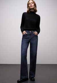 Granatowe jeansy o szerokich nogawkach wykonane z denimu, zestawione z czarnym swetrem z wysokim kołnierzem. Strój ma luźny fason z przednimi kieszeniami.