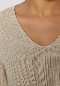 Gros plan sur la poitrine d'une personne portant un pull beige à col en V tricoté, montrant le tissu texturé et le contraste avec le ton de la peau.