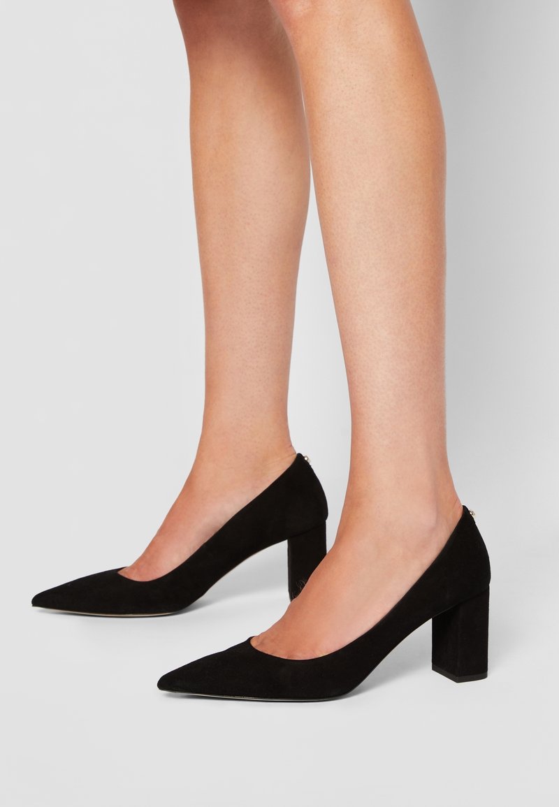 Jambes portant des chaussures en daim noir à bout pointu et talon bloc sur un fond clair uni.