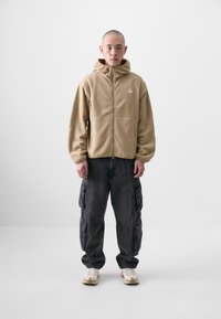 Beige Fleecejacke mit Reißverschluss und Kapuze, kombiniert mit dunkelgrauen Cargo-Shorts mit Taschen. Das Model trägt hellfarbene Sneaker.