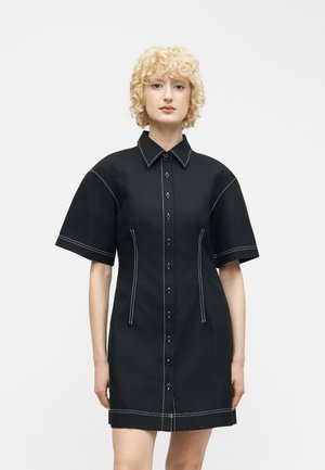 Lyshåret kvinde med krøllet hår iført en sort kortærmet knap-op kjole med hvide syninger på en ensfarvet hvid baggrund.