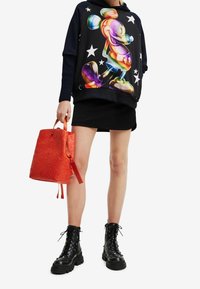 Sudadera negra con capucha con un gráfico colorido de Mickey Mouse y estrellas blancas, combinada con un bolso tipo balde texturizado en naranja y botas de combate negras.