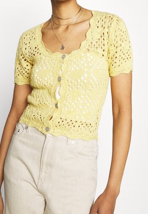 Kobieta ubrana w żółty krótki sweter na guziki, do tego jasnobeżowe spodnie z wysokim stanem i wielowarstwowe srebrne naszyjniki.