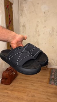 Χέρι κρατάει μαύρα σανδάλια με σταυρωτά λουριά μέσα σε ένα δωμάτιο με ξύλινο πάτωμα, ένα μοτίβο χαλί και μια δερμάτινη τσάντα.