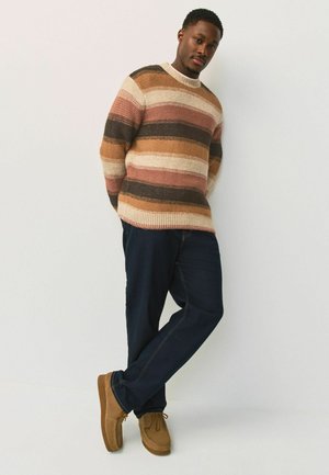 Gestreifter Strickpullover in den Farbtönen Pink, Braun und Creme, kombiniert mit dunkelblauer Jeans und braunen Wildleder-Mokassins.