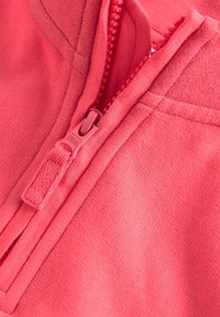 Gros plan sur la fermeture éclair partiellement fermée d'un sweat à capuche rouge, avec une languette en tissu et des coutures visibles sur un tissu doux.
