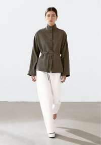 Vrouw die wandelt in een donkerbruin langmouwtop met riem en staande kraag, met witte wijde pantalon en witte schoenen, neutrale achtergrond.