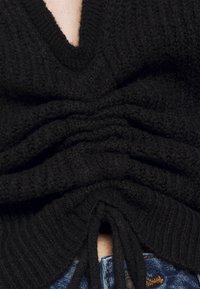 Svart stickad tröja med V-ringning, som har samlad textur och justerbara snören i botten. Bärs över blå jeans.