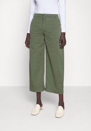 Woman wearing high-waisted, wide-leg olive green cropped pants, white long-sleeve top, and cream slip-on shoes, standing on light background.