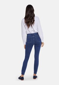 Jeans ajustados de color azul oscuro hechos de denim con una textura suave. Presentan una cinturilla alta, bolsillos traseros y piernas ajustadas. Combinados con una camisa blanca.