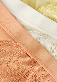 Drie stoffen stukken met kanten randen in perzik, wit en geel. Elk heeft een gladde textuur met ingewikkelde kantpatronen en zichtbare stiksels.