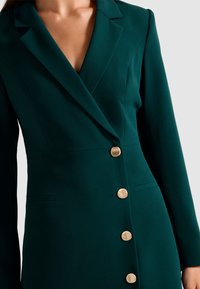 Donna che indossa un blazer dress su misura di colore verde scuro con bottoni dorati e scollatura a V profonda su uno sfondo chiaro.