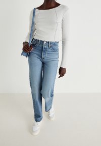 Top blanco de manga larga con relieve y bordes festoneados, combinado con jeans de tiro alto azul claro y zapatillas blancas. Modelo sosteniendo un bolso azul.