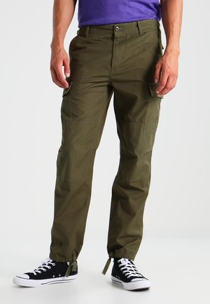 Mężczyzna w oliwkowozielonych spodniach typu cargo z elastycznymi mankietami i czarnych wysokich trampkach, stojący na jednolitym tle.