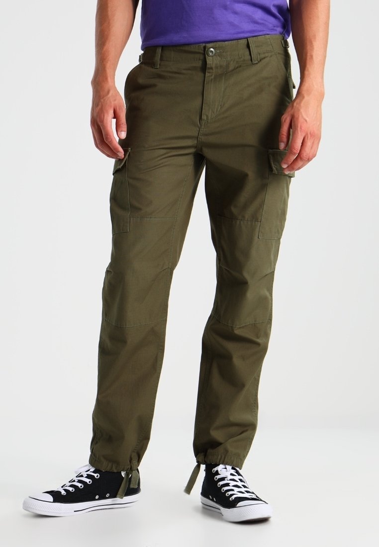 Mann trägt olivgrüne Cargo-Hosen mit elastischen Bündchen und schwarze High-Top-Sneaker, steht vor einem einfarbigen Hintergrund.