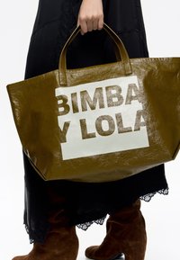 Olive zelena torba iz sijočega materiala, z veliko belo ploščo in bold črnim napisom "BIMBA Y LOLA." Ima trde ročaje.