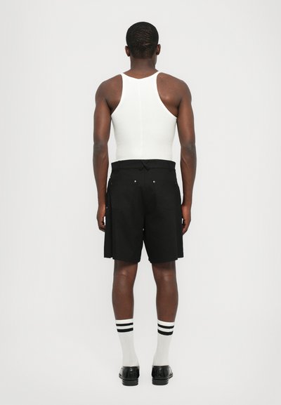 Schwarze Shorts mit einem lässigen Schnitt, kombiniert mit einem weißen Tanktop und weißen Socken mit schwarzen Streifen, von hinten betrachtet.