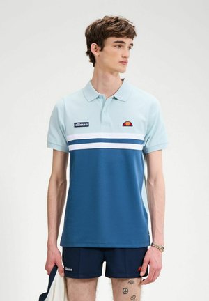 Giovane uomo che indossa una polo ellesse a blocchi di colore azzurro e blu navy e pantaloncini blu navy, tiene in mano sinistra un panno bianco.
