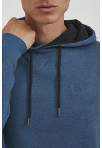 Blå hættetrøje lavet af struktureret stof med et kontrasterende sort indre lag. Har snore og minimal logo-detajle på brystet.