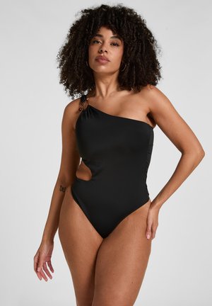 Frau mit lockigem Haar, die einen schwarzen Badeanzug mit einer Schulter und einem seitlichen Ausschnitt trägt, steht mit einer Hand in der Hüfte vor einem einfarbigen Hintergrund.