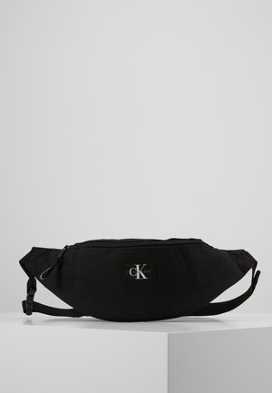 Czarna materiałowa nerka o zakrzywionym kształcie, zapinana na zamek, z regulowanym paskiem i naszywką z logo na przodzie. Widoczna faktura powierzchni.