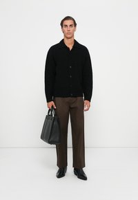 Sort strikket cardigan med krave, knaplukning, båret over brune bukser, holdende en slank sort læder tote taske.