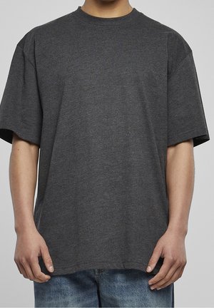 Hombre vestido con una camiseta de manga corta gris oscuro y holgada, y jeans azules, contra un fondo gris liso.