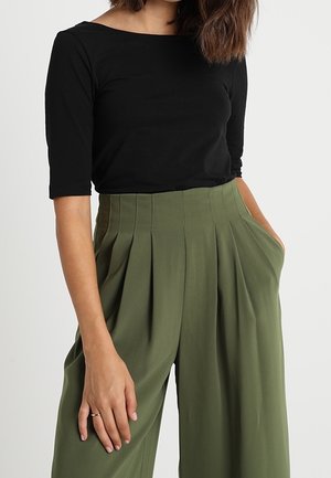 Vrouw draagt een zwart topje met driekwart mouwen en olijfgroene, wijde broek met hoge taille, plooien en zakken.