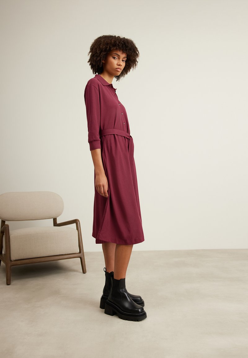 Bordeauxfarbendes Hemdkleid mit Kragen, Knopfleiste und Bindegürtel; Dreiviertelärmel; kombiniert mit klobigen schwarzen Knöchelstiefeln.