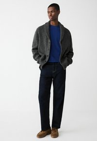 Gray knitted cardigan with a shawl collar, navy blue crew neck shirt, dark jeans, and tan moccasin shoes. Simple button detailing on cardigan.