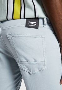Pantalon en denim bleu clair avec une poche à rabat, des coutures décoratives et une étiquette noire avec un logo de ciseaux au niveau de la taille.
