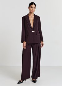 Abito su misura bordeaux con blazer a profondo scollo a V, dettagli in oro, pantaloni a gamba larga e eleganti tacchi neri. Tessuto dalla superficie liscia.