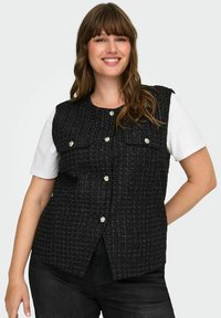 Črna suknjič iz tvida z teksturiranim vzorcem, sprednjim zapiranjem na gumbe in dvema prsni žepoma. Nošen preko bele srajce.