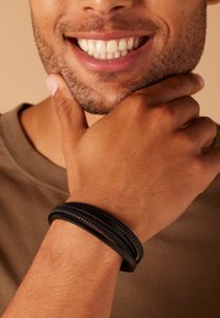 Zwart gevlochten leren armband gedragen om een hand met een gemiddelde huidskleur. De persoon heeft een lichtbruine shirt aan en een goed verzorgde baard.