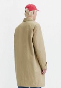 Cappotto beige con una vestibilità comoda, maniche lunghe e chiusura a un bottone. Presenta tasche laterali e una superficie liscia. Indossato con un cappello rosso.