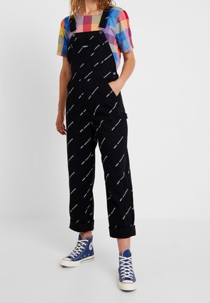 Peto negro con estampado de texto blanco por todo el tejido, tirantes en los hombros, bolsillos frontales, puños remangados, combinado con una camisa de cuadros multicolor y zapatillas azules.