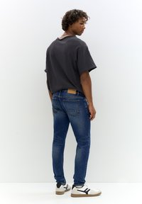 Szürke, túlméretezett póló rövid ujjakkal, sötétkék, slim-fit farmerral és fekete akcentusú fehér sportcipővel párosítva. Barna bőr folt a farmereken.