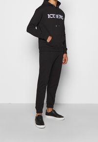 Sudadera negra con logo blanco "ICEBERG", bolsillo frontal y capucha con cordones. Combinada con pantalones de chándal negros a juego y zapatillas negras.
