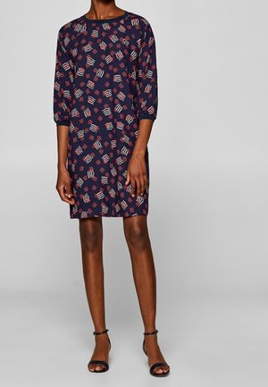 Femme portant une robe bleu marine longueur genou avec des motifs abstraits rouges et blancs et des manches trois-quarts, associée à des sandales noires à brides à la cheville.