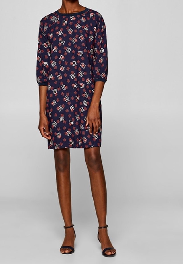 Femme portant une robe bleu marine longueur genou avec des motifs abstraits rouges et blancs et des manches trois-quarts, associée à des sandales noires à brides à la cheville.