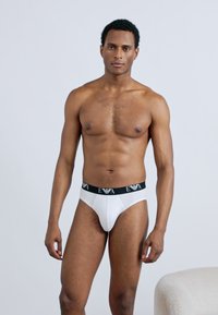 Slip da uomo bianchi con fascia in vita nera EWA, caratterizzati da un design aderente e una texture liscia. Modello in piedi che mostra la vista frontale su uno sfondo neutro.