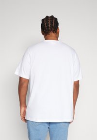 Camiseta de algodón blanca con un ajuste relajado, mangas cortas y escote redondo, mostrada desde la parte de atrás. Jeans de mezclilla parcialmente visibles.