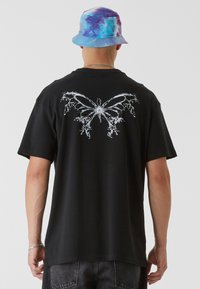 T-shirt noir en coton présentant un grand graphique de papillon blanc dans le dos. Associé à un chapeau seau multicolore en tie-dye.