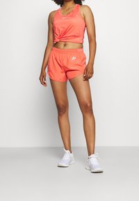 Top deportivo y pantalones cortos Nike en color coral, fabricados con un tejido ligero y de diseño ajustado, con un logo en la parte frontal, combinados con zapatillas deportivas blancas.