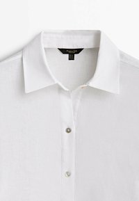 Camicia bianca in lino con colletto classico, due bottoni visibili e etichetta nera del marchio all'interno del collo.
